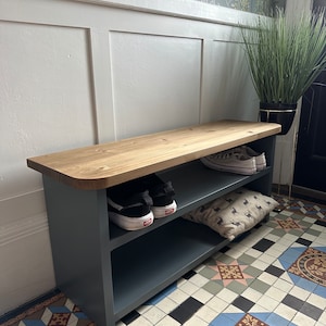May include: A wooden shoe storage bench with a grey base and a natural wood top. The bench has two shelves, holding shoes and a cushion. The bench is against a white wall with floral wallpaper.