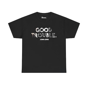 May include: Black t-shirt with the words "GOOD TROUBLE." in a grayscale font. Below the text is "JOHN LEWIS" in a smaller font. The shirt has a crew neck and short sleeves. The brand name "THE RESISTANCE." is printed on the back of the neck.