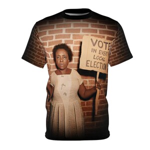 May include: A black crew neck t-shirt featuring a vintage photograph of a woman holding a sign that reads "VOTE IN EVERY LOCAL ELECTION". The image is set against a brick wall background.
