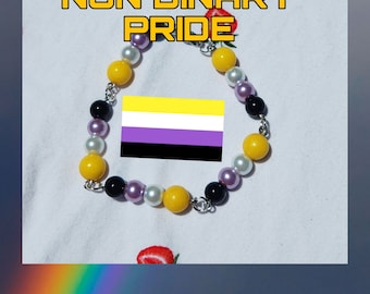 May include: A bracelet with yellow, black, white, and purple beads. The non-binary pride flag is featured, along with the words "NON BINARY PRIDE" in yellow block letters.