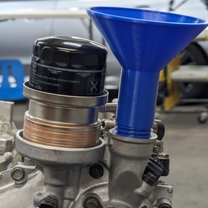 May include: A blue plastic funnel attached to a silver engine part. The funnel is used to pour oil into the engine. The engine part has a black oil filter with the text "CAUTION: TURN OIL FILTER AFTER CONTACT GASKET.  TIGHTEN TO ADJUST LEVEL. BLACK LTD." printed on it.