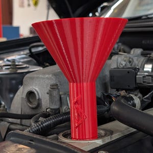 May include: A bright red plastic funnel with a wide, conical top and a cylindrical neck featuring a raised geometric logo. The funnel is inserted into the oil filler port of a car engine, showing visible 3D printed layer lines on its surface.