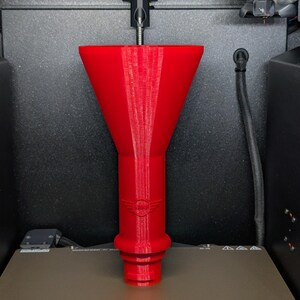 May include: A bright red, 3D-printed funnel with a wide top and a narrow, cylindrical base. The funnel has a logo near the base and is sitting on a gray surface. The image is taken inside a 3D printer.