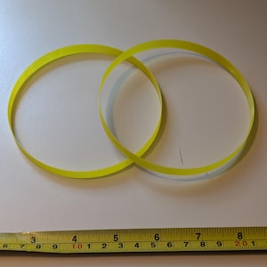 以下が含まれることがあります： 2つの重なり合った明るい黄色の円形バンド。 画像の下部には、センチメートル単位の測定値を持つ定規があります。 バンドは薄く柔軟な素材でできているようです。 バンドは白い表面にあります。