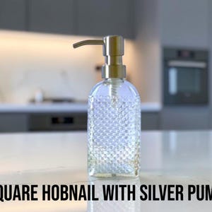 May include: A clear glass soap dispenser with a square hobnail texture and a metallic silver pump mechanism. The bottle features a distinct raised dot pattern across its surface and is topped with a functional silver-colored dispensing nozzle.