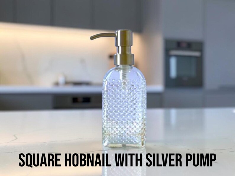 May include: A clear glass soap dispenser with a square hobnail texture and a metallic silver pump mechanism. The bottle features a distinct raised dot pattern across its surface and is topped with a functional silver-colored dispensing nozzle.