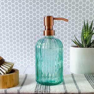May include: A clear, light green glass soap dispenser with vertical ribbed texture and a metallic copper-colored pump mechanism. The cylindrical bottle features a rounded base and a slender neck, topped with a functional pump for dispensing liquid soap.