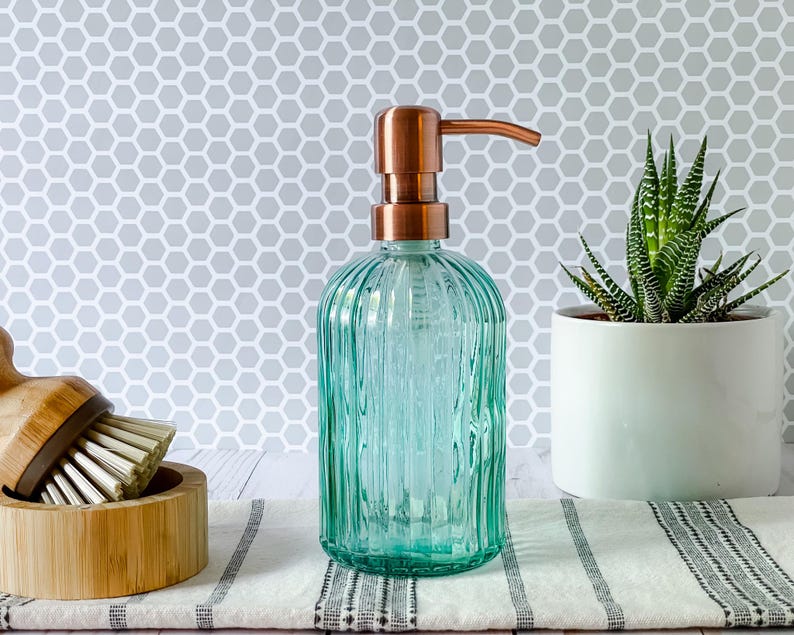 May include: A clear, light green glass soap dispenser with vertical ribbed texture and a metallic copper-colored pump mechanism. The cylindrical bottle features a rounded base and a slender neck, topped with a functional pump for dispensing liquid soap.