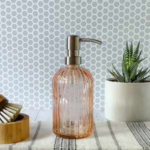 May include: A light pink, ribbed glass soap dispenser with a brushed silver metal pump mechanism. The cylindrical bottle features vertical fluting throughout its body, providing a textured appearance for this functional bathroom or kitchen accessory.