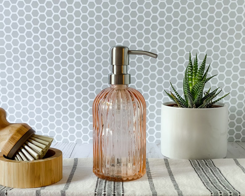 May include: A light pink, ribbed glass soap dispenser with a brushed silver metal pump mechanism. The cylindrical bottle features vertical fluting throughout its body, providing a textured appearance for this functional bathroom or kitchen accessory.