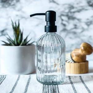 May include: A clear glass soap dispenser with a vertical ribbed texture and a matte black pump mechanism. The cylindrical bottle features a rounded base and a slender neck, topped with a functional black plastic pump for dispensing liquid soap.