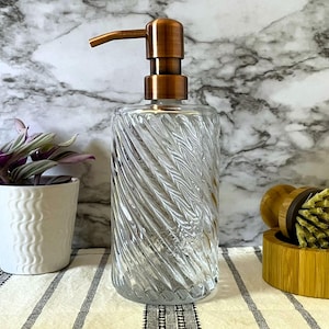 May include: A clear glass soap dispenser with a copper pump top. The dispenser has a swirl design and is sitting on a white and black striped surface.