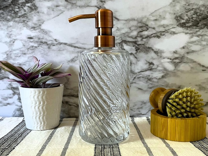 May include: A clear glass soap dispenser with a copper pump top. The dispenser has a swirl design and is sitting on a white and black striped surface.