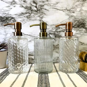 May include: Three glass soap dispensers with copper pumps. The dispensers have different textured glass designs. The dispensers are on a white and black striped surface.