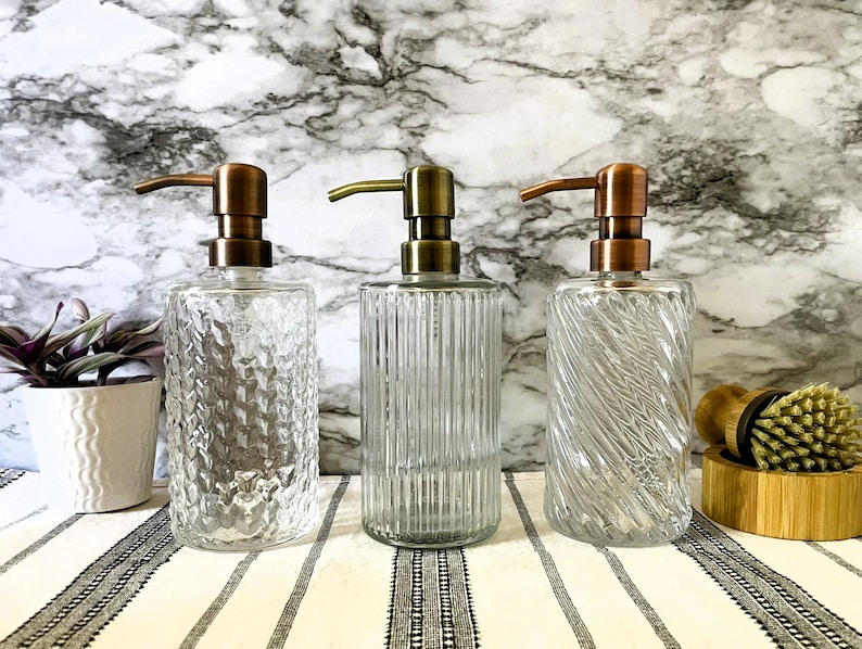May include: Three glass soap dispensers with copper pumps. The dispensers have different textured glass designs. The dispensers are on a white and black striped surface.