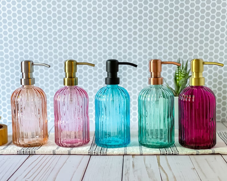 May include: Five glass soap dispensers with vertical ribbing and pump mechanisms. The dispensers are colored light amber, pink, bright blue, teal, and deep magenta, each topped with a metallic pump in silver, gold, black, copper, or brass.