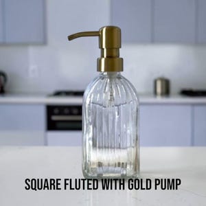 May include: A clear glass soap dispenser with a square base and vertical fluted detailing, topped with a metallic gold pump mechanism. The text "SQUARE FLUTED WITH GOLD PUMP" is printed in black at the bottom of the image.