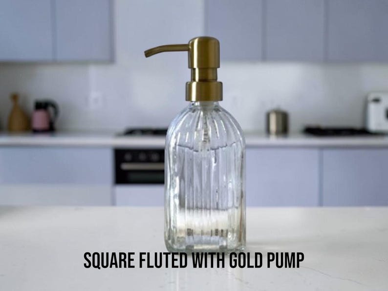 May include: A clear glass soap dispenser with a square base and vertical fluted detailing, topped with a metallic gold pump mechanism. The text "SQUARE FLUTED WITH GOLD PUMP" is printed in black at the bottom of the image.