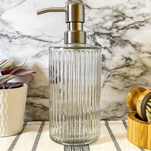 May include: A clear glass soap dispenser with a stainless steel pump and a ribbed design. The dispenser is sitting on a white and black striped surface.
