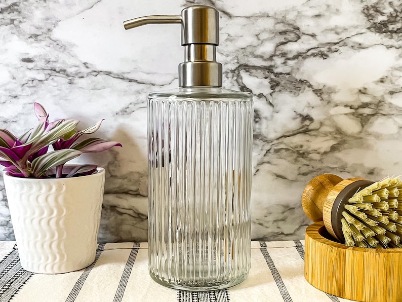 May include: A clear glass soap dispenser with a stainless steel pump and a ribbed design. The dispenser is sitting on a white and black striped surface.