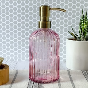 May include: A pink glass soap dispenser with a vertical ribbed texture and a metallic brass-toned pump mechanism. The cylindrical bottle features a rounded base and a slender neck, topped with a functional manual push-down dispenser nozzle.