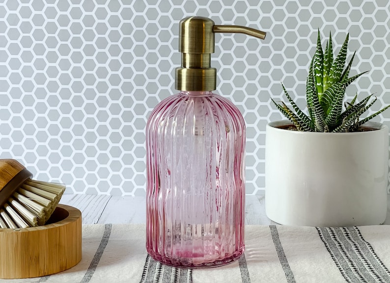 May include: A pink glass soap dispenser with a vertical ribbed texture and a metallic brass-toned pump mechanism. The cylindrical bottle features a rounded base and a slender neck, topped with a functional manual push-down dispenser nozzle.