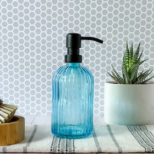May include: A translucent light blue glass soap dispenser with a vertical ribbed texture and a matte black pump mechanism. The cylindrical bottle features a rounded shoulder and a flat base, designed for dispensing liquid soap or lotion.