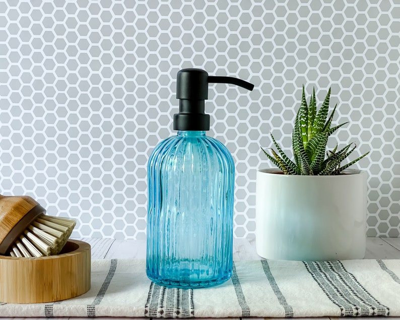May include: A translucent light blue glass soap dispenser with a vertical ribbed texture and a matte black pump mechanism. The cylindrical bottle features a rounded shoulder and a flat base, designed for dispensing liquid soap or lotion.