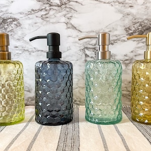 May include: Four decorative glass soap dispensers with honeycomb patterns. The dispensers are in shades of green, blue, and yellow, each with a different colored pump. The dispensers are arranged on a white and gray striped surface.