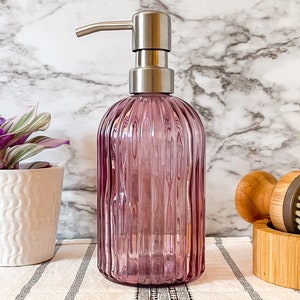 May include: A translucent purple glass soap dispenser with a vertical ribbed texture and a brushed silver metal pump mechanism. The cylindrical bottle features a rounded base and a narrow neck, topped with a functional stainless steel pump head.