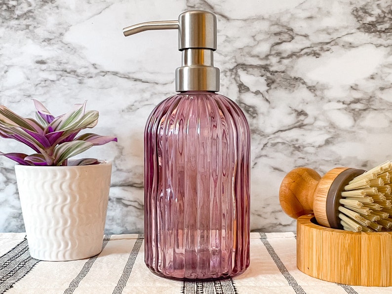 May include: A translucent purple glass soap dispenser with a vertical ribbed texture and a brushed silver metal pump mechanism. The cylindrical bottle features a rounded base and a narrow neck, topped with a functional stainless steel pump head.