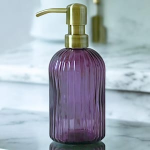 May include: A purple glass soap dispenser with a vertical ribbed texture and a brushed brass pump mechanism. The cylindrical bottle features a rounded base and a metallic gold-toned top that extends into a curved spout for dispensing liquid soap.