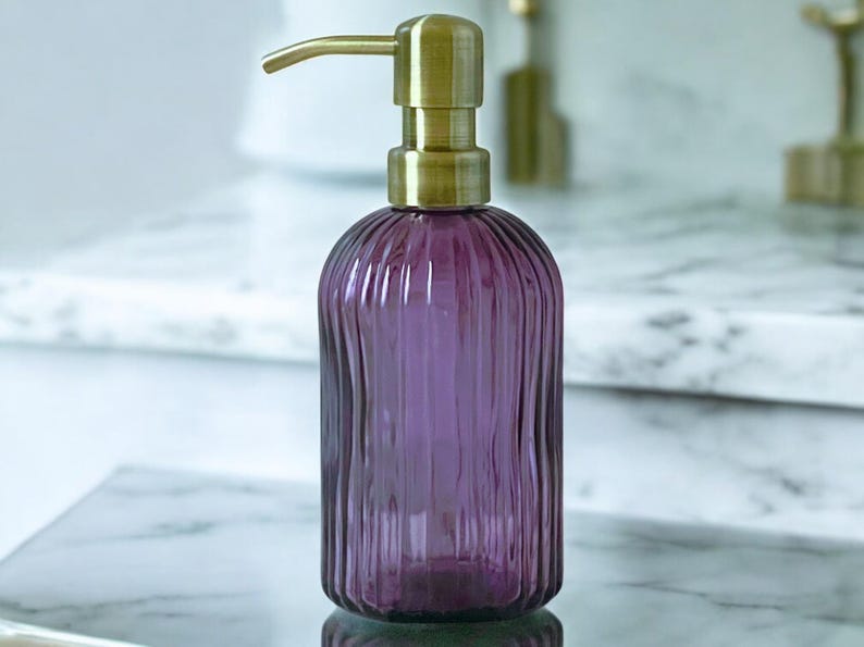 May include: A purple glass soap dispenser with a vertical ribbed texture and a brushed brass pump mechanism. The cylindrical bottle features a rounded base and a metallic gold-toned top that extends into a curved spout for dispensing liquid soap.