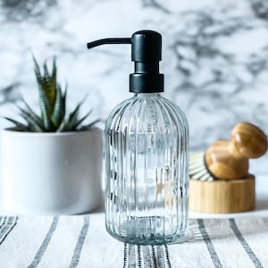 May include: A clear glass soap dispenser with a vertical ribbed texture and a matte black pump mechanism. The bottle has a rounded base and a cylindrical body, designed for dispensing liquid soap or lotion in a bathroom or kitchen setting.