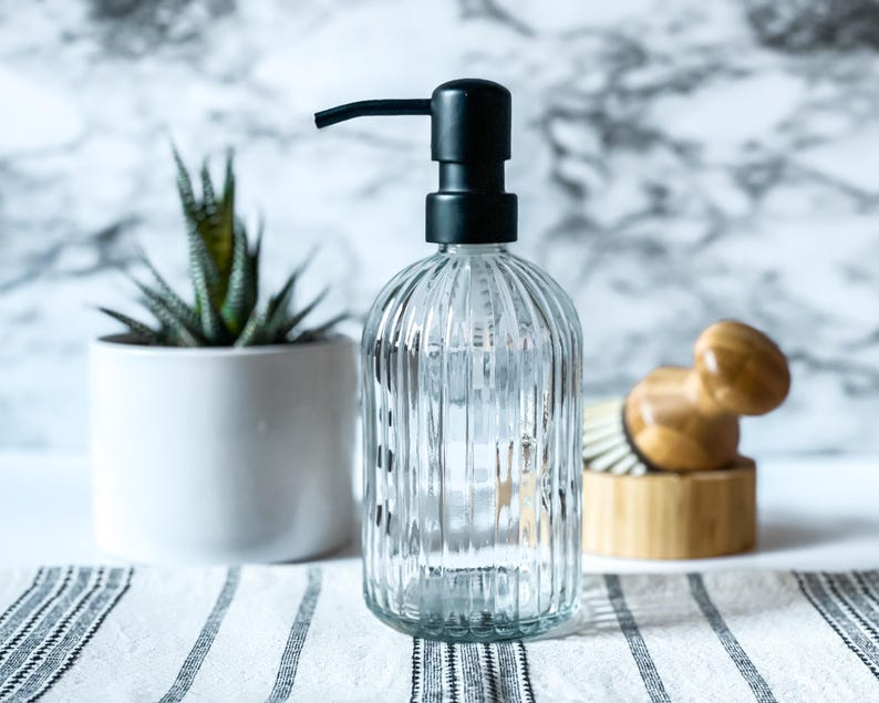 May include: A clear glass soap dispenser with a vertical ribbed texture and a matte black pump mechanism. The bottle has a rounded base and a cylindrical body, designed for dispensing liquid soap or lotion in a bathroom or kitchen setting.