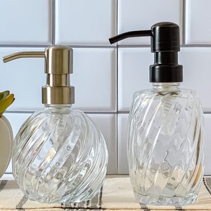 May include: Two clear glass soap dispensers with a swirl design. The dispenser on the left has a silver pump and the dispenser on the right has a black pump.