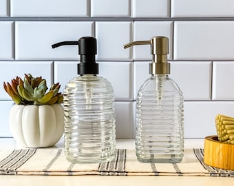 May include: Two clear glass soap dispensers with ribbed designs. One dispenser has a black pump and the other has a silver pump. Both dispensers are sitting on a striped towel.