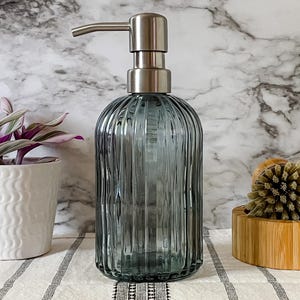May include: A translucent, ribbed, blue-gray glass soap dispenser with a brushed silver-tone metal pump mechanism. The cylindrical bottle features vertical fluting throughout its body, providing a textured appearance for this bathroom accessory.