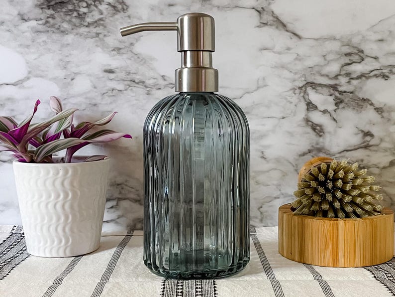 May include: A translucent, ribbed, blue-gray glass soap dispenser with a brushed silver-tone metal pump mechanism. The cylindrical bottle features vertical fluting throughout its body, providing a textured appearance for this bathroom accessory.