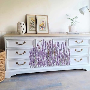 K&ouml;nnte beinhalten: Wei&szlig;e Kommode mit sechs Schubladen, die auf der Vorderseite mit einem Lavendel-Blumendesign verziert ist. Die Kommode hat goldfarbene Griffe und eine Holzplatte.