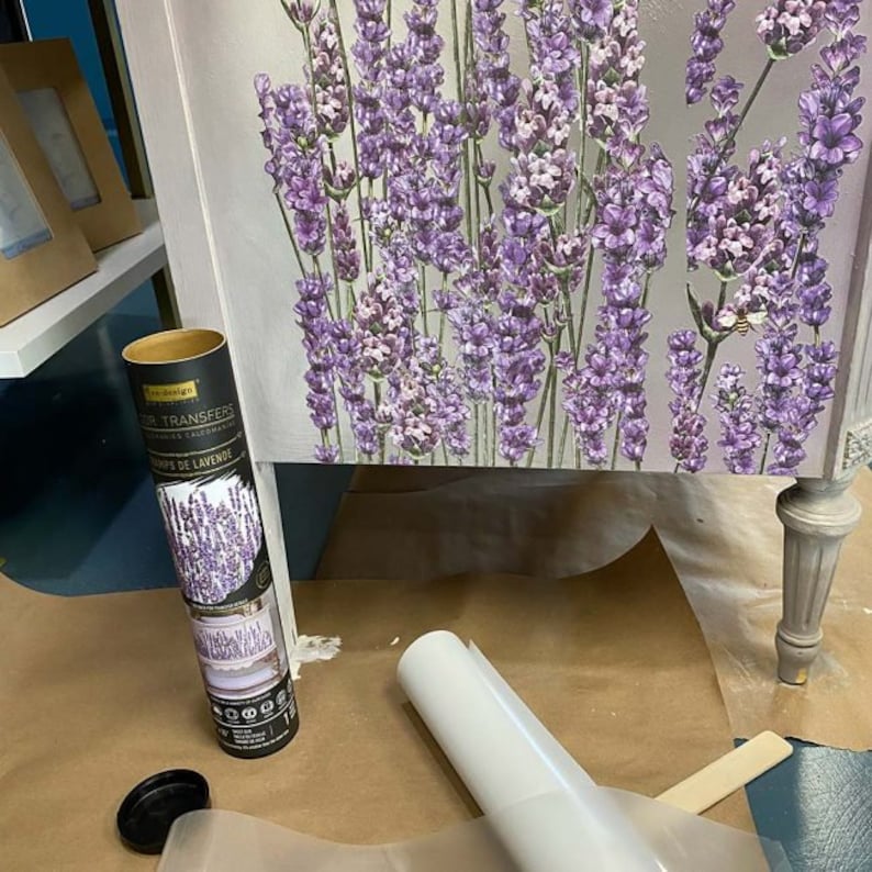 K&ouml;nnte beinhalten: Eine wei&szlig;e Kommode mit einem Lavendel-Blumenmuster. Die Kommode hat eine Schublade und ein Regal. Die Schublade ist offen und auf dem Regal liegt eine Rolle Transferpapier. Das Transferpapier ist mit "Decoupage Transfers" und "Bouquets de Lavande" beschriftet.