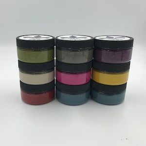 Pu&ograve; includere: Un set di 6 piccoli barattoli di vernice in vari colori, tra cui verde, grigio, viola, beige, rosa, giallo, rosso, blu e blu-verde. I barattoli sono impilati in tre colonne di due barattoli ciascuna. Ogni barattolo ha un coperchio nero con un design a giglio.
