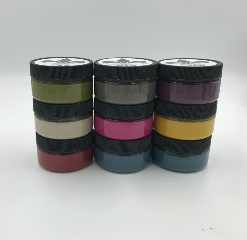 Pu&ograve; includere: Un set di 6 piccoli barattoli di vernice in vari colori, tra cui verde, grigio, viola, beige, rosa, giallo, rosso, blu e blu-verde. I barattoli sono impilati in tre colonne di due barattoli ciascuna. Ogni barattolo ha un coperchio nero con un design a giglio.