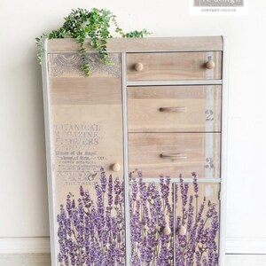 K&ouml;nnte beinhalten: Ein Holzschr&auml;nkchen mit vier Schubladen und zwei T&uuml;ren. Der Schrank ist wei&szlig; lackiert und hat eine Used-Look-Oberfl&auml;che. Die T&uuml;ren sind mit einem Lavendel-Blumenmuster und dem Text "BOTANICAL MAGAZINE FLOWERS PHANTWOR" verziert.