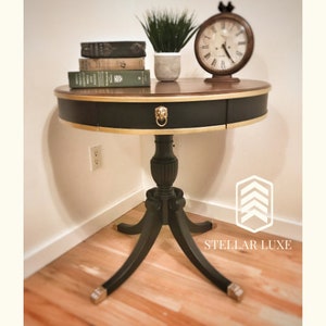 May include: A black and gold round end table with a single drawer and three legs. The table has a lion's head pull on the drawer. The table is in a corner of a room with a clock and a potted plant on top.