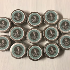 May include: A collection of 12 small, round, silver tins with labels featuring a stylized owl and the text "Wise Owl Furniture Salve". Each tin has a different scent name printed on the label.
