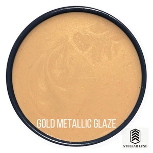 May include: A close-up of a small container of gold metallic glaze. The glaze is a shimmery, metallic gold color. The container is labeled "GOLD METALLIC GLAZE" and "STELLAR LUXE".