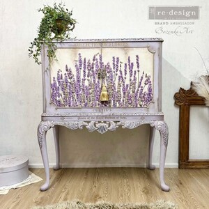 K&ouml;nnte beinhalten: Ein lavendelfarbenes Schr&auml;nkchen mit einem floralen Design auf der Vorderseite. Das Schr&auml;nkchen hat eine Glast&uuml;r und einen dekorativen Quasten. Das Schr&auml;nkchen steht auf einem Tisch mit verzierten Beinen.