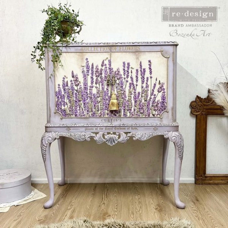 K&ouml;nnte beinhalten: Ein lavendelfarbenes Schr&auml;nkchen mit einem floralen Design auf der Vorderseite. Das Schr&auml;nkchen hat eine Glast&uuml;r und einen dekorativen Quasten. Das Schr&auml;nkchen steht auf einem Tisch mit verzierten Beinen.