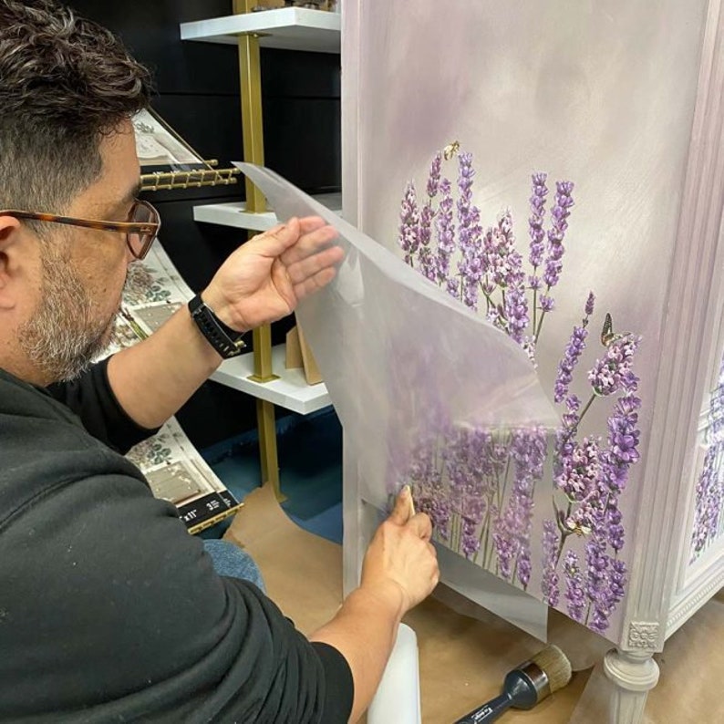 K&ouml;nnte beinhalten: Eine Person bringt eine transparente Schutzfolie auf einen in Lavendelfarben bemalten Schrank an. Der Schrank hat einen hellvioletten Hintergrund mit einem floralen Design aus Lavendelbl&uuml;ten und -bl&auml;ttern. Der Schrank hat eine wei&szlig;e, abgenutzte Oberfl&auml;che und goldene Akzente.